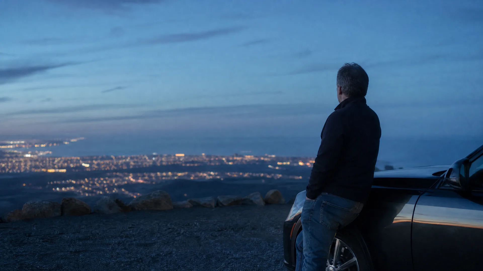 A man in a dark jacket and jeans leans against a car at a gravel overlook at dusk, facing a valley of city lights below.